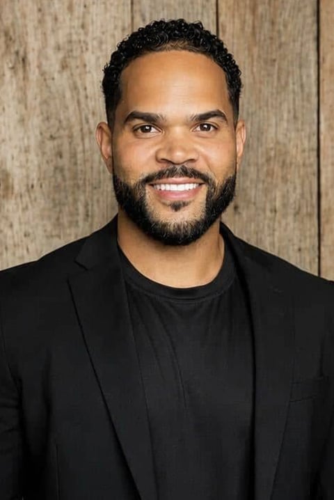 Professional headshot of a smiling man with dark curly hair and beard, wearing a black blazer over a black shirt against a wooden background.